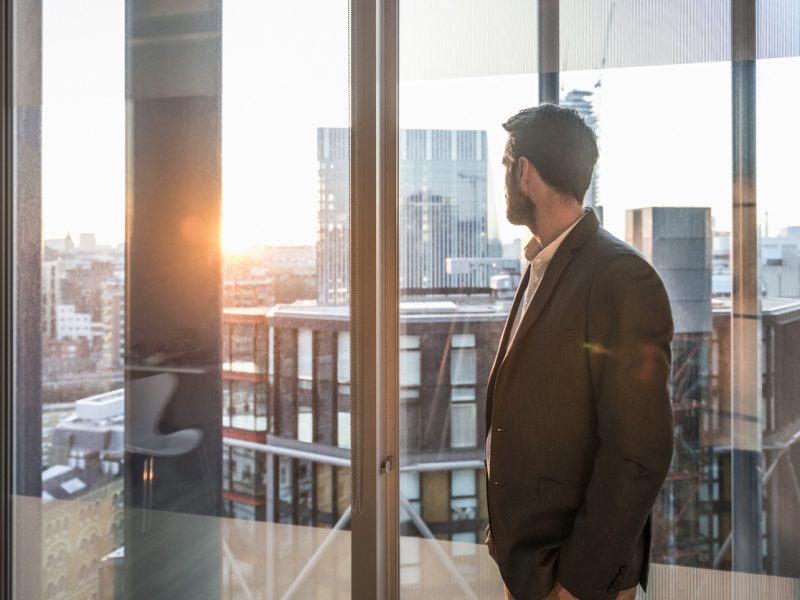 A confident executive in a business suit reflects at sunrise through a high-rise window, symbolizing clarity and momentum after a career break.