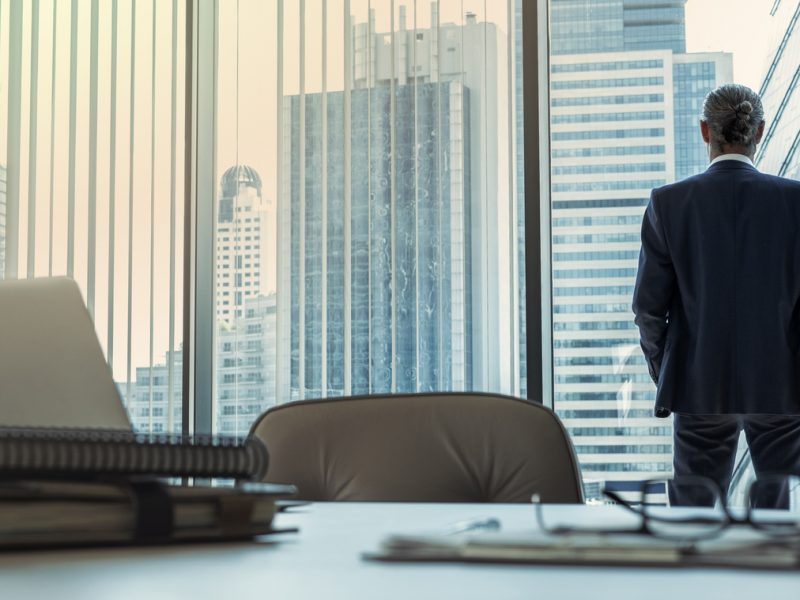 Executive in suit overlooking city skyline through office window, symbolizing career reflection and transition.