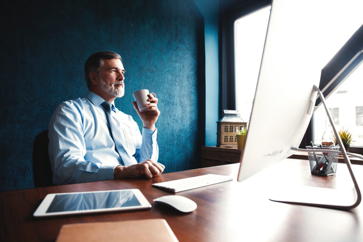 Mature executive man in shirt and tie working remotely at a desk, drinking coffee and reflecting by the window – symbolizing leadership with balance.