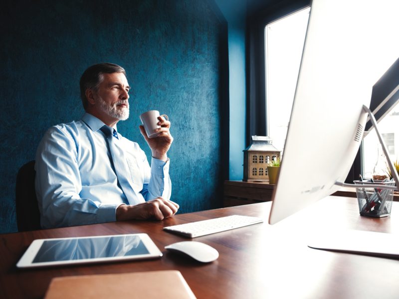 Mature executive man in shirt and tie working remotely at a desk, drinking coffee and reflecting by the window – symbolizing leadership with balance.