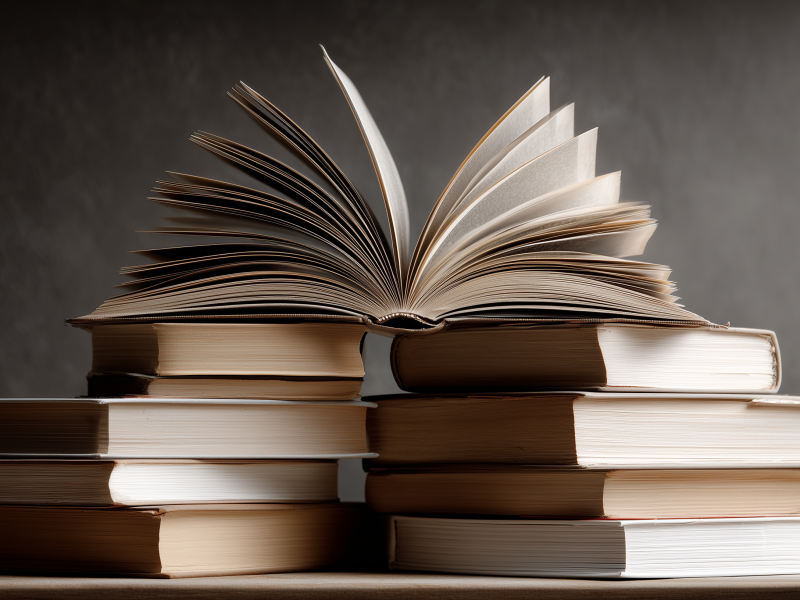 Stack of hardcover books with one open in the middle, pages fanned out, on a wooden table against a dark background.